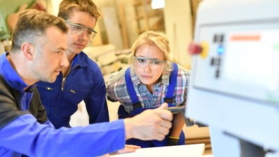 Zu sehen sind zwei Auszubildende in einem Industriebetrieb, die einen Vorgang an einer Maschine erklärt bekommen.