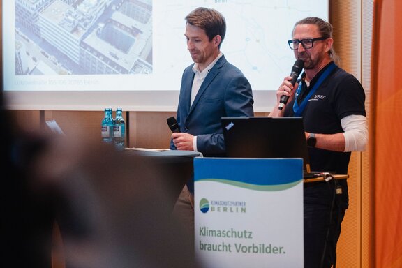 Zu sehen sind zwei Männer auf einer Bühne, einer hält ein Mikrofon und spricht zum Publikum.
