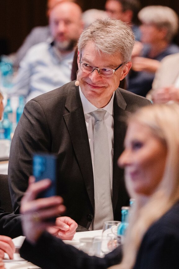 Zu sehen ist ein älterer Herr mit Brille und grauen Haaren. Er trägt einen Anzug, unscharf im Vordergrund nimmt eine blonde Frau gerade ein Foto mit ihrem handy auf.