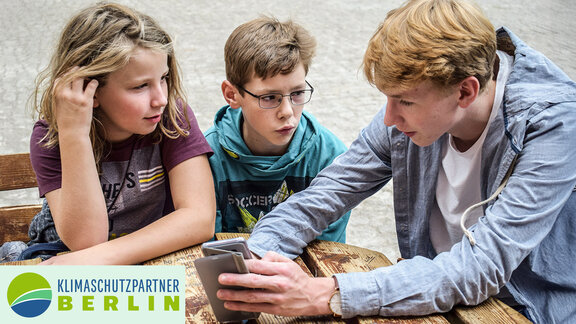 Ein Jugendlicher sitzt mit zwei anderen Kindern an einem Tisch und erklärt etwas an einem Handy. 