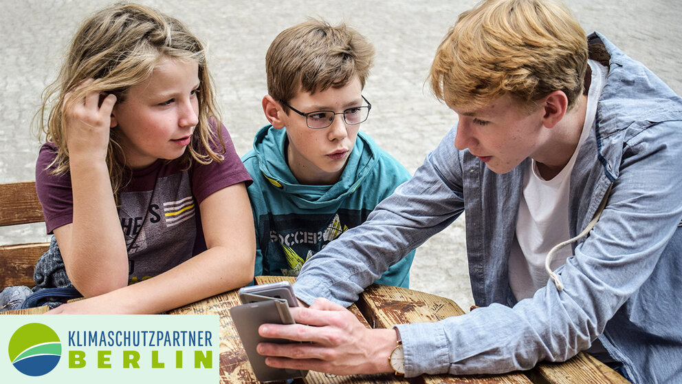 Ein Jugendlicher sitzt mit zwei anderen Kindern an einem Tisch und erklärt etwas an einem Handy. 
