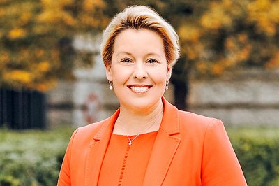 Frau in orangefarbenem Blazer und Kleid steht vor einer Hecke und einem Gebäude aus Stein.
