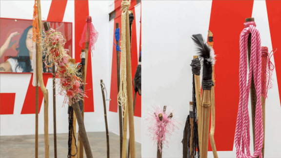 Detailansicht einer Kunstinstallation mit mehreren Stäben, die mit verschiedenen Materialien wie bunten Fäden, Federn und Seilen verziert sind. Im Hintergrund sind rote Wände sichtbar.
