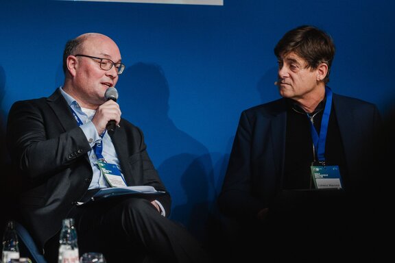 Zwei Männer unterhalten sich sitzend auf einer Bühne. Der gerade Sprechende hält ein Mikrofon.