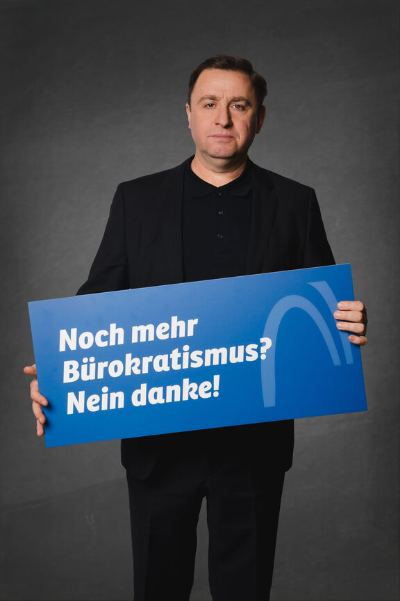 Eine Person steht vor einem neutralen grauen Hintergrund und hält ein großes blaues Schild mit der weißen Aufschrift: "Noch mehr Bürokratismus? Nein danke!"