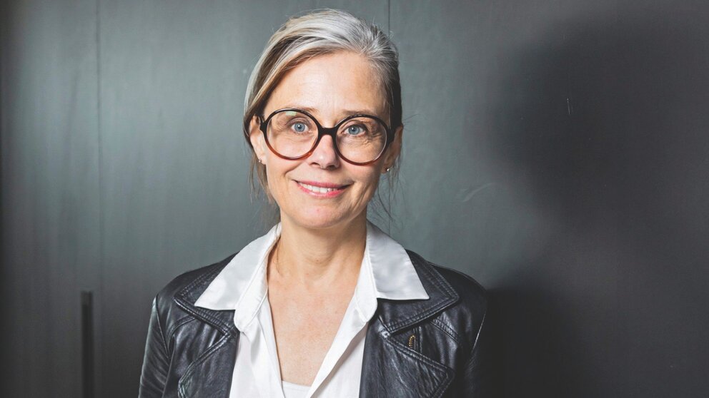 Zu sehen ist eine Frau mittleren Alters mit hellen Haaren, Brille, weißer Bluse und schwarzer Lederjacke.