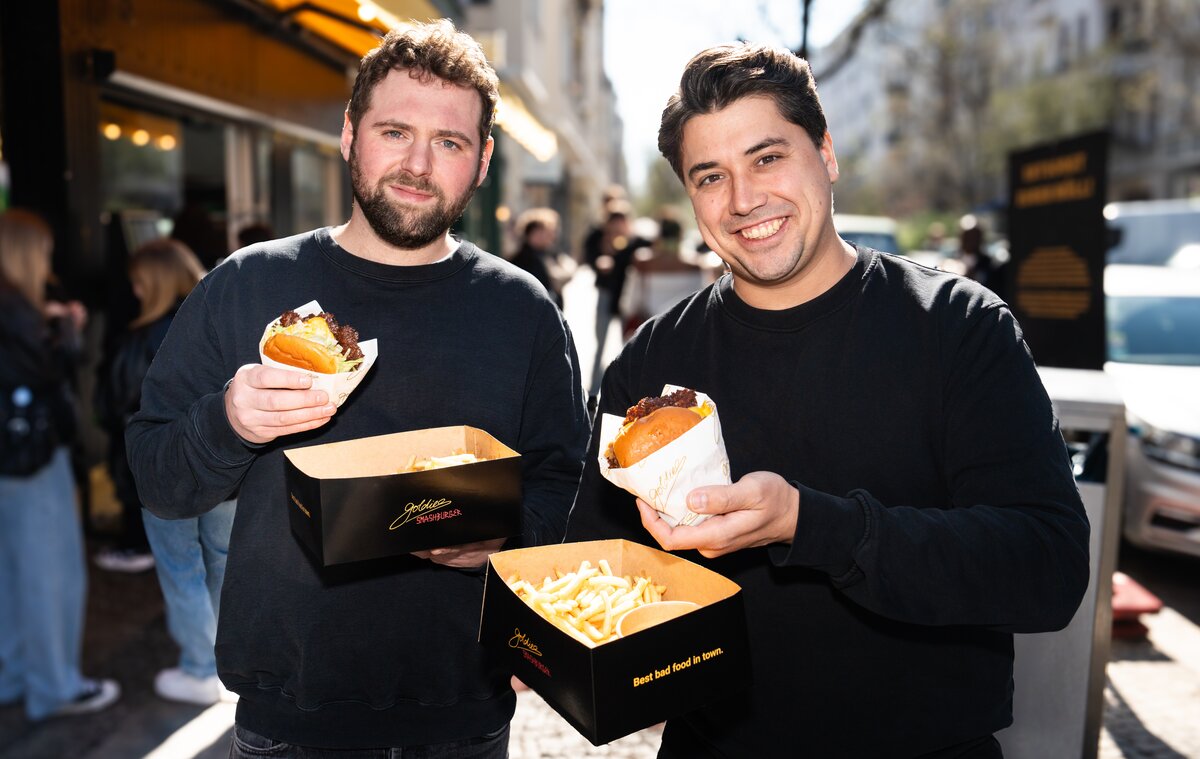 Zu sehen sind zwei Männer, die mit Fast Food in der Hand posieren – es sind die Gründer von Goldies.