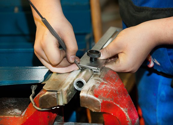 Hände messen mit einem Messschieber einen Metallzylinder, der in einem Schraubstock eingespannt ist