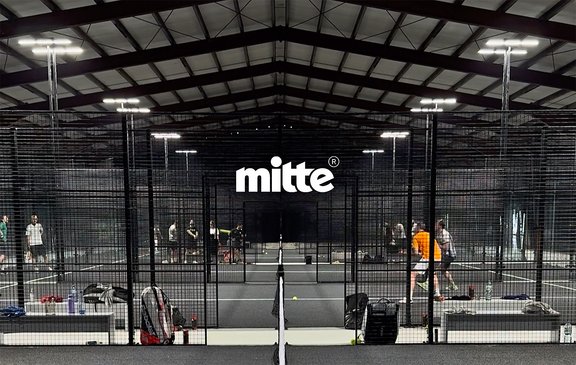 Innenaufnahme einer Halle mit mehreren Padel-Tennisplätzen, Spielern und Ausrüstung, mittig das weiße Logo 'mitte'.