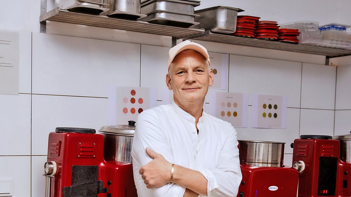 Person in weißer Kochjacke steht mit verschränkten Armen in einer professionellen Küche vor roten Küchenmaschinen und Metallbehältern.