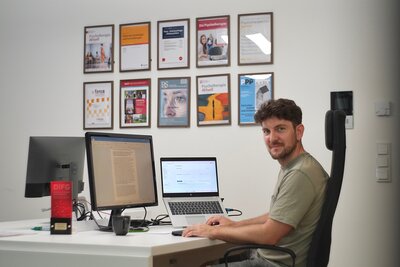 Ein Mann sitzt vor einem Laptop an einem Schreibtisch. Auf dem Schreibtisch steht ebenfalls ein Bildschirm. An der Wand dahinter hängen mehrere Auszeichnungen und Bilder 