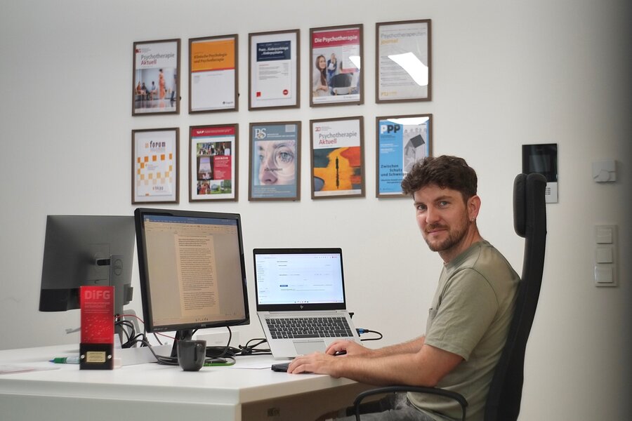 Ein Mann sitzt vor einem Laptop an einem Schreibtisch. Auf dem Schreibtisch steht ebenfalls ein Bildschirm. An der Wand dahinter hängen mehrere Auszeichnungen und Bilder 