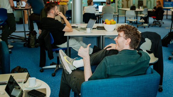 Einblicke in den KI-Hackathon in Berlin im Oktober 2026. Ein junger Mann sitzt in einem blauen Sessel und macht den Daumen nach oben.