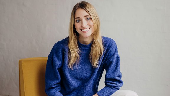 Auf dem Bild ist eine blonde Frau mit blauem Puulover zu sehen, die auf einem Stuhl sitzt.