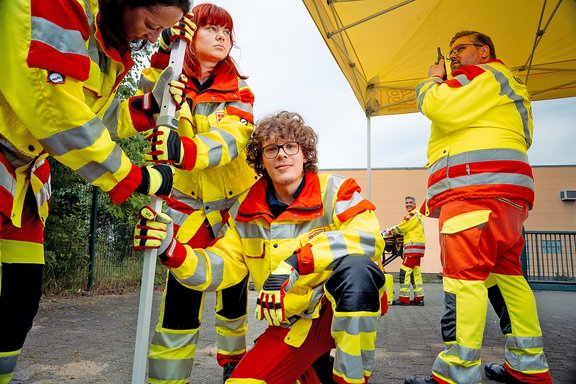 Fünf Rettungskräfte in gelb-roten Schutzanzügen bei einer Übung im Freien unter einem gelben Zelt, eine Person kniet und hält ein Gerät mit Stange.