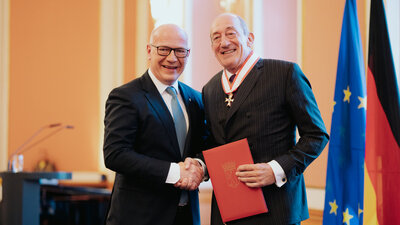 Zu sehen sind zwei Männer in dunklen Anzügen, die vor zwei Fahnen sich die Hand schütteln, es sind Jan Eder und Kai Wegner