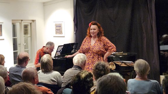 Frau in rotem gemustertem Kleid steht vor einem Flügelklavier, umgeben von sitzendem Publikum in einem kleinen Raum mit weißen Wänden und Fenstern.