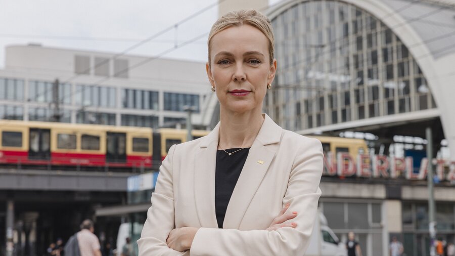 Zu sehen ist eine Frau mit blonden Haaren und einem hellen Sakko. Sie steht mit verschränkten Armen vor der Berliner Friedrichstraße. Es handelt sich um Manja Schreiner.