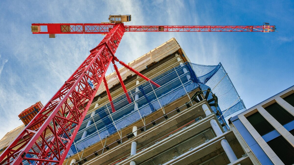 Von unten ist ein Rohbau eines sich im Bau befindenden Wohnhauses zu sehen, welches sich über mehrere Etagen erstreckt. Unmittelbar vor dem Haus steht ein großer roter Baukran. Auf der Hälfte der Höhe des Gebäudes befindet sich ein blaues Netz um das Gebäude gespannt, welches herunterfallende Baumaterialien abfangen kann.