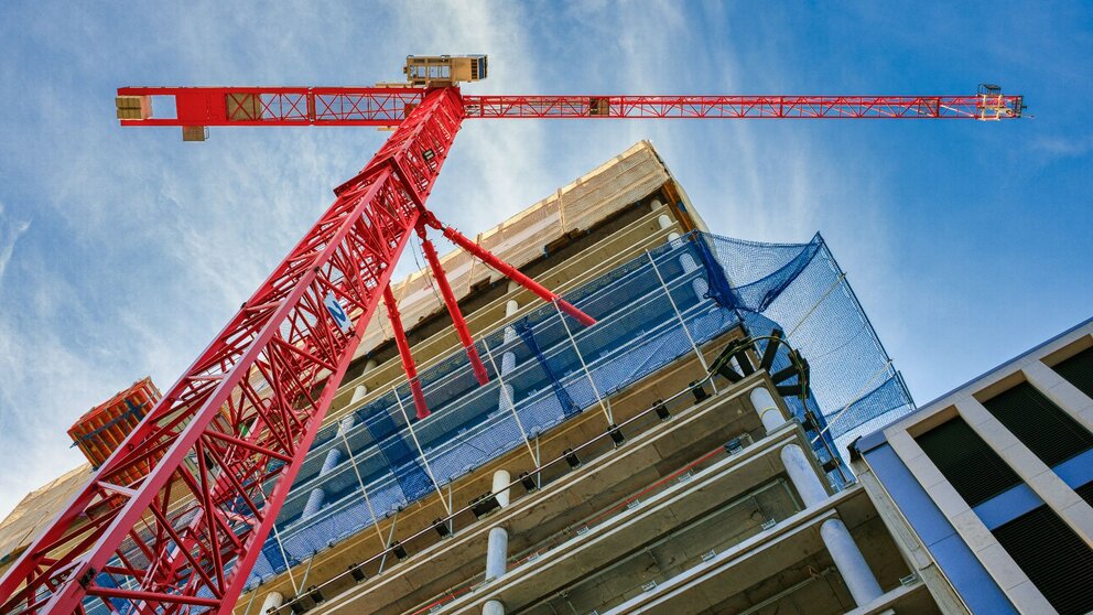 Von unten ist ein Rohbau eines sich im Bau befindenden Wohnhauses zu sehen, welches sich über mehrere Etagen erstreckt. Unmittelbar vor dem Haus steht ein großer roter Baukran. Auf der Hälfte der Höhe des Gebäudes befindet sich ein blaues Netz um das Gebäude gespannt, welches herunterfallende Baumaterialien abfangen kann.