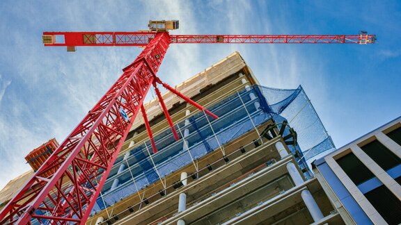 Von unten ist ein Rohbau eines sich im Bau befindenden Wohnhauses zu sehen, welches sich über mehrere Etagen erstreckt. Unmittelbar vor dem Haus steht ein großer roter Baukran. Auf der Hälfte der Höhe des Gebäudes befindet sich ein blaues Netz um das Gebäude gespannt, welches herunterfallende Baumaterialien abfangen kann.