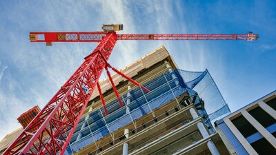 Von unten ist ein Rohbau eines sich im Bau befindenden Wohnhauses zu sehen, welches sich über mehrere Etagen erstreckt. Unmittelbar vor dem Haus steht ein großer roter Baukran. Auf der Hälfte der Höhe des Gebäudes befindet sich ein blaues Netz um das Gebäude gespannt, welches herunterfallende Baumaterialien abfangen kann.