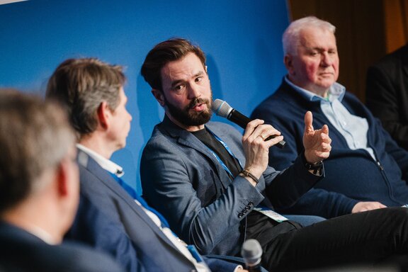 Vier Männer sitzen auf einer Bühne, einer spricht in einem Mikrofon zu den anderen.