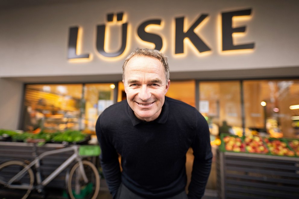 Mann mit grauen Haaren trägt schwarzen Pullover, steht vor einem Geschäft mit dem beleuchteten Schriftzug 'LÜSKE' und Obst- und Gemüseständen im Hintergrund.
