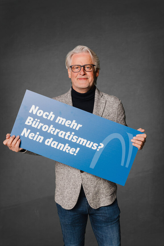 Eine Person steht vor einem neutralen grauen Hintergrund und hält ein großes blaues Schild mit der weißen Aufschrift: "Noch mehr Bürokratismus? Nein danke!"
