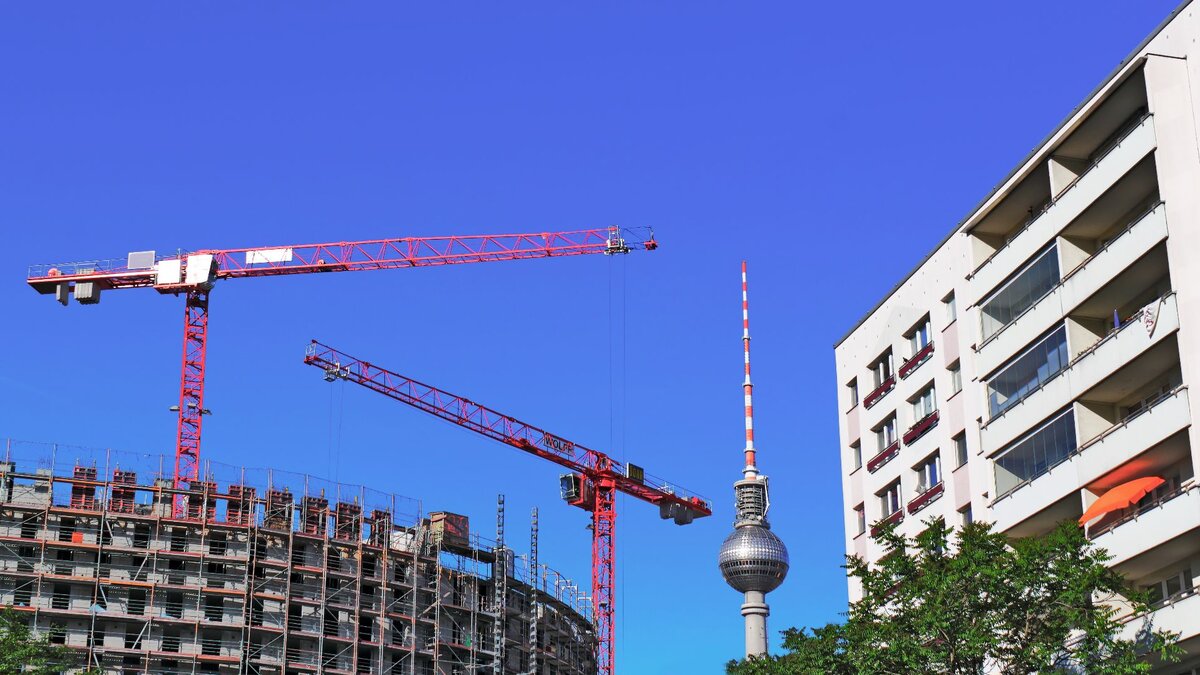 Zu sehen ist ein Wohnhaus, ein Turm sowie ein sich im Bau befindliches Wohnhaus, hinter welchem zwei rote Baukräne stehen. Es handelt sich um eine Szene in Berlin mit dem Fernsehturm im Hintergrund.