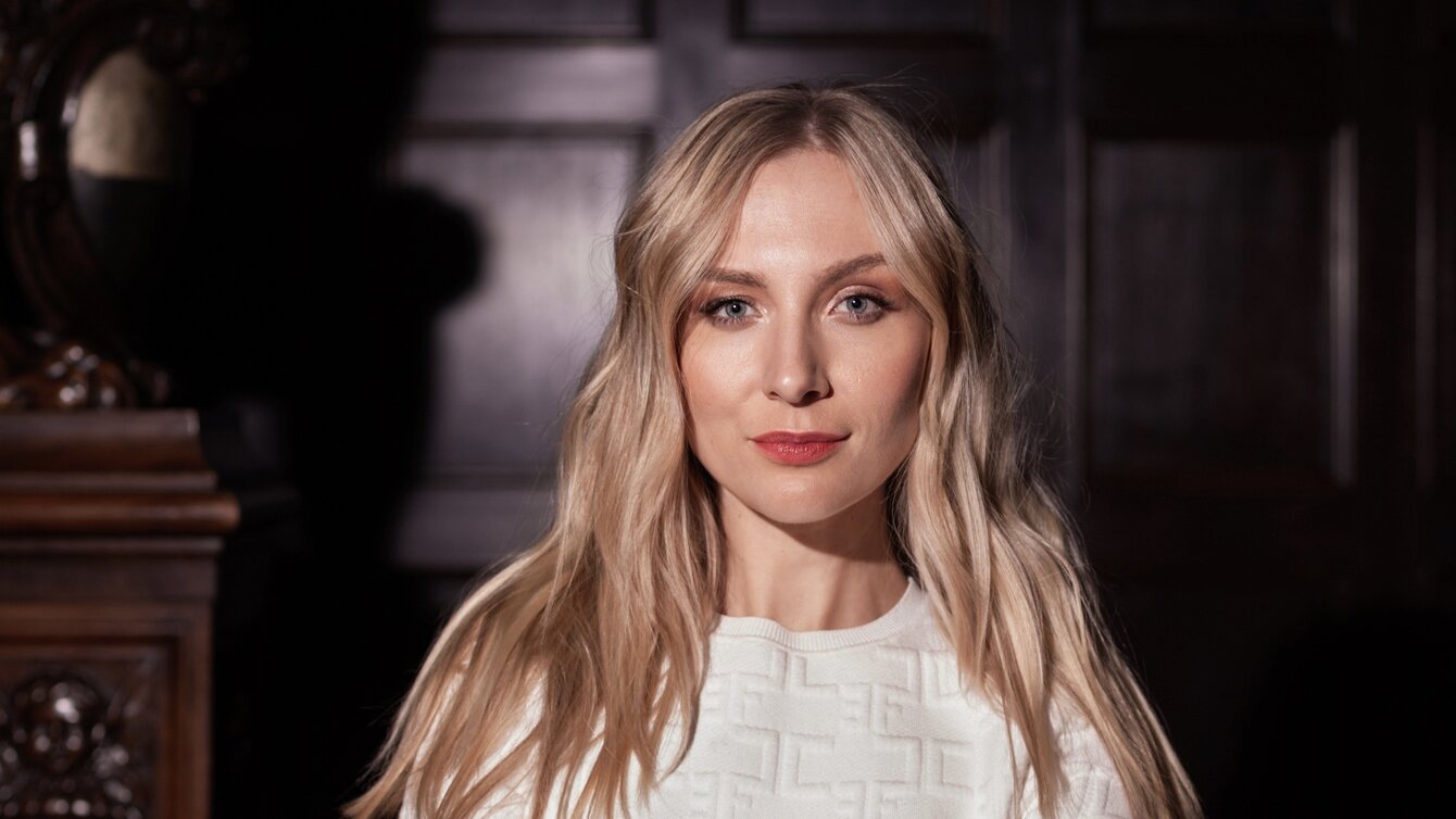 Zu sehen ist eine junge blonde Frau, die in die Kamera schaut. Sie trägt ein weißes Oberteil.