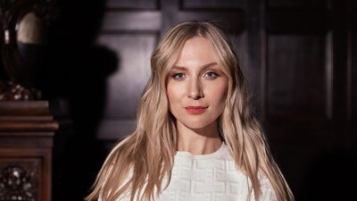 Zu sehen ist eine junge blonde Frau, die in die Kamera schaut. Sie trägt ein weißes Oberteil.