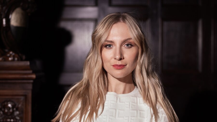 Zu sehen ist eine junge blonde Frau, die in die Kamera schaut. Sie trägt ein weißes Oberteil.