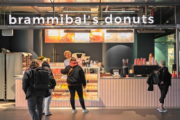 Donutladen 'brammibals donuts' mit beleuchtetem Schriftzug, Kunden vor Verkaufstresen mit ausgestellten Donuts und Menütafeln
