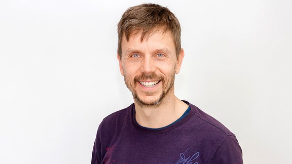 Mann mit braunen Haaren trägt ein dunkelviolettes T-Shirt mit blauem Muster vor weißem Hintergrund.