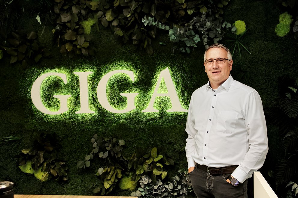 Mann in weißem Hemd steht vor einer Wand mit leuchtendem Schriftzug 'GIGA' und grünen Pflanzen