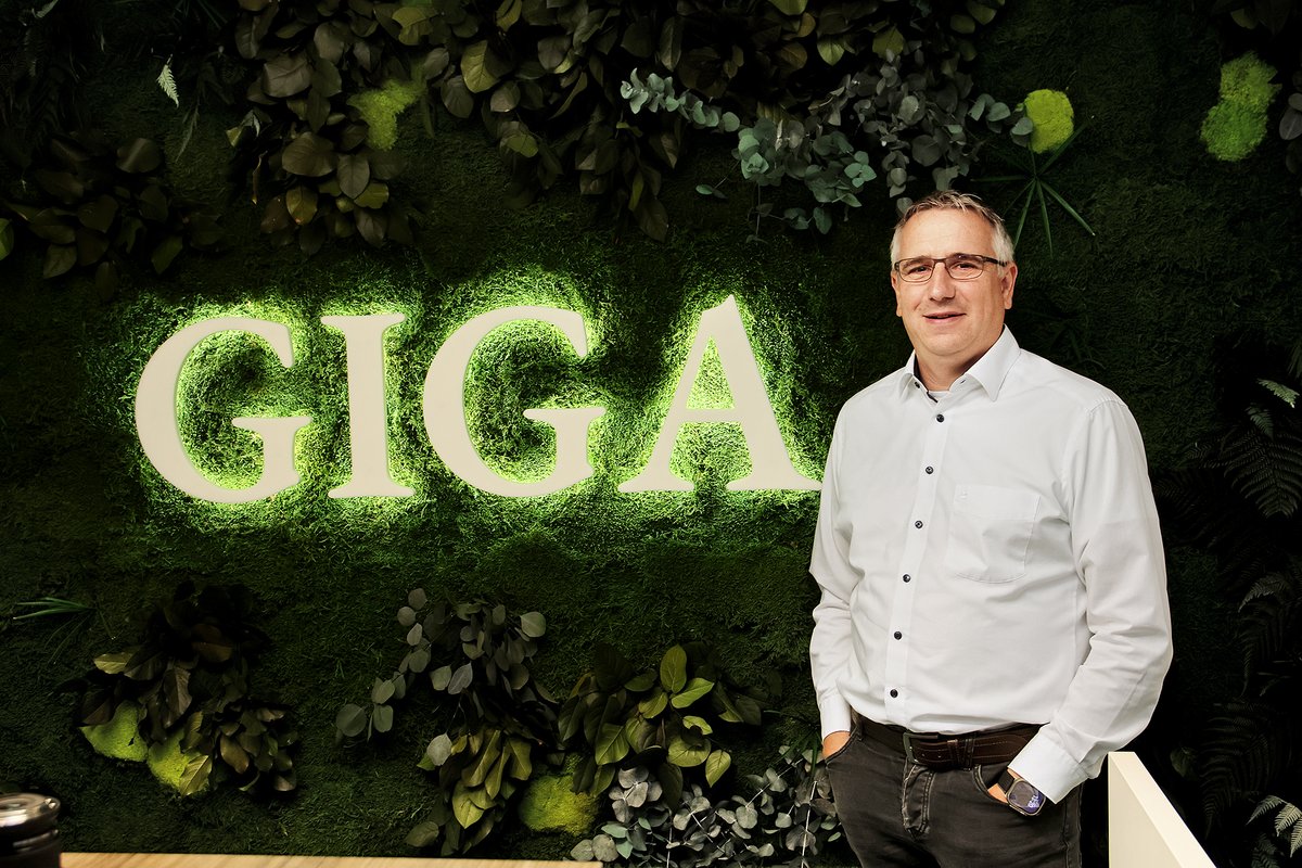 Mann in weißem Hemd steht vor einer Wand mit leuchtendem Schriftzug 'GIGA' und grünen Pflanzen
