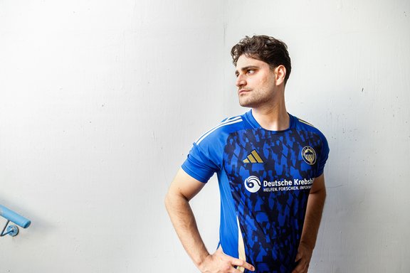 Junger Mann in blauem Sporttrikot mit Logo der Deutschen Krebshilfe, steht vor weißer Wand und schaut nach rechts