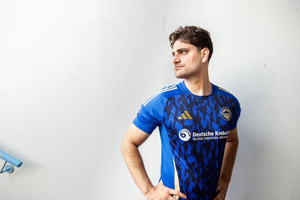 Junger Mann in blauem Sporttrikot mit Logo der Deutschen Krebshilfe, steht vor weißer Wand und schaut nach rechts