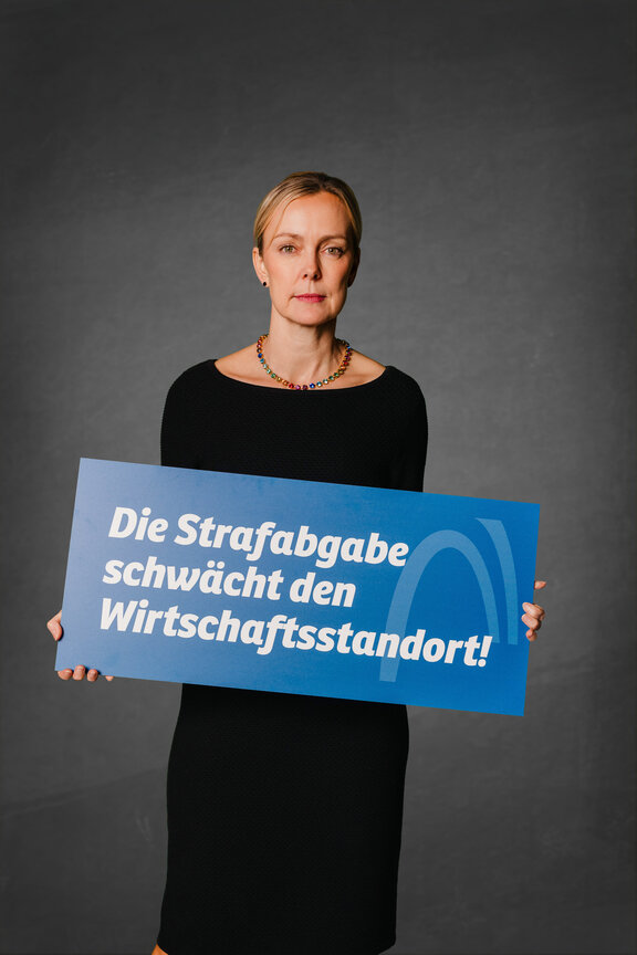 Eine Person steht vor einem neutralen grauen Hintergrund und hält ein großes blaues Schild mit der weißen Aufschrift: "Die Strafabgabe schwächt den Wirtschaftsstandort!"