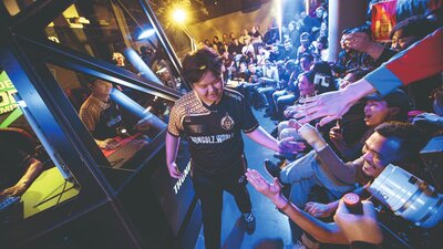 Ein Teilnehmer eines E-Sport-Wettbewerbs läuft am Publikum vorbei und klatscht deren Hände ab. Im Hintergrund sieht man eine Kabine, in der andere Wettbewerber gamen. 