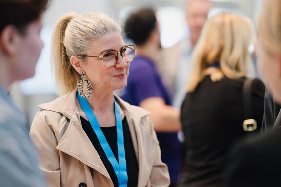 Zu sehen ist eine blonde Frau mit Brille im Gespräch mit anderen Personen.