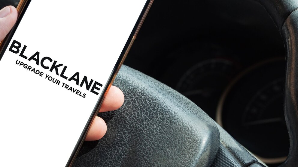 Zu sehen ist ein Smartphone in der Hand einer Person. Die Hand liegt auf einem Autolenkrad. Auf dem Smartphone steht "Blacklane". Dabei handelt es sich um den Berliner Chauffeurdienstleister.