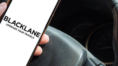 Zu sehen ist ein Smartphone in der Hand einer Person. Die Hand liegt auf einem Autolenkrad. Auf dem Smartphone steht "Blacklane". Dabei handelt es sich um den Berliner Chauffeurdienstleister.