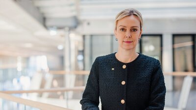 Zu sehen ist eine Frau mit blonden Haaren und einer schwarzen Jacke, die neutral in die Kamera schaut