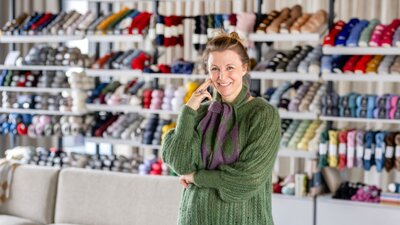Zu sehen ist eine Frau in grünem Pullover, die vor einem Regal steht in dem zahllose bunte Stoffspulen hängen.