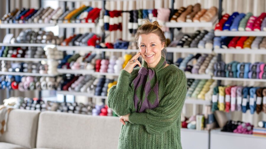 Zu sehen ist eine Frau in grünem Pullover, die vor einem Regal steht in dem zahllose bunte Stoffspulen hängen.