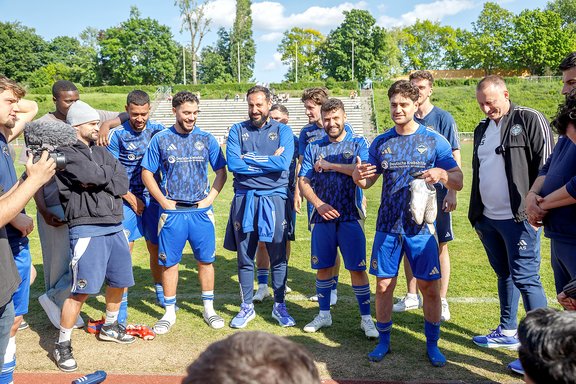 Fußballspieler in blauen Trikots und Trainer stehen im Kreis auf dem Spielfeld und hören zu, einige lächeln