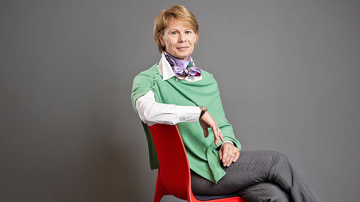 Person sitzt seitlich auf einem roten Stuhl, trägt graue Hose, weiße Bluse und grünen Pullover, hält Arm auf Stuhllehne.