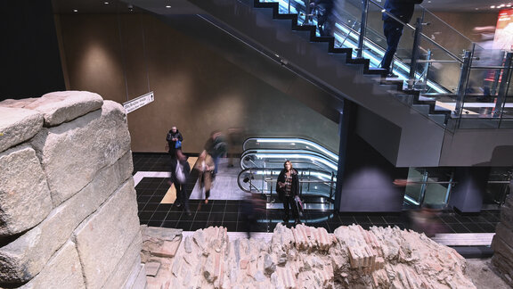 Eine Frau schaut auf eine antike Mauer. Sie steht in einer modernen U-Bahn-Station mit vielen Passanten. 